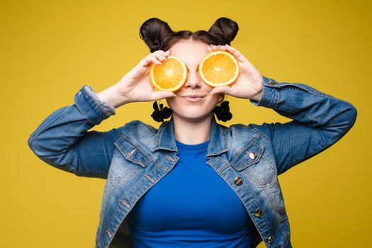 Studio headshot of young funny brunette with hairstyle and red lips holding halved oranges on eyes against bright yellow background.