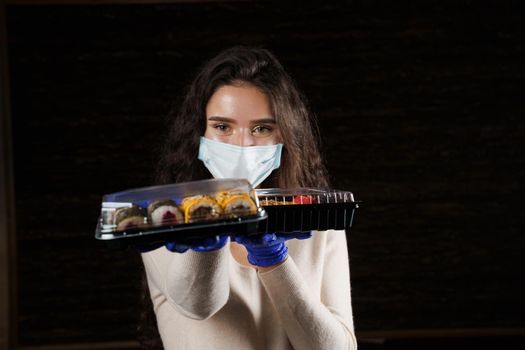 Girl sushi courier in medical mask with 2 boxes. Sushi set in box healthy food delivery service by car. Japanese cuisine: rolls, soy sauce, wasabi.