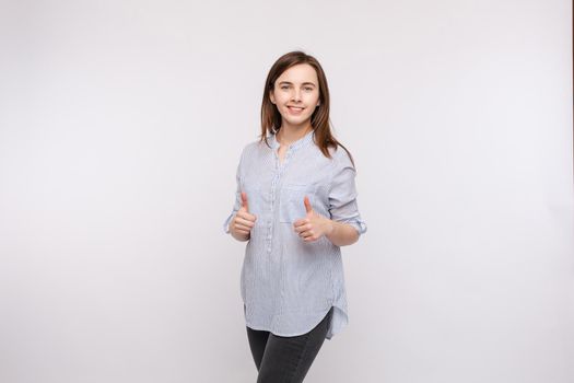 Studio portrait of pretty brunette woman in shirt showing thumbs up and smiling at camera with pleasure and satisfaction. Isolate on white.