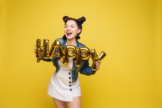 Attractive happy woman laughing holding big gold letters posing at yellow studio background medium long shot. Beautiful fashionable young girl relaxing having positive emotion