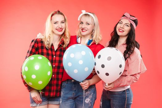 Stock photo of three cheerful beautiful and stylish girls hugging and smiling at camera. They are holding colorful dotted air balloons. Isolate on red background.