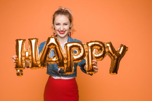Stock photo portrait of attractive girl with red lips holding shining bright inflatable word HAPPY. Isolate on orange background.