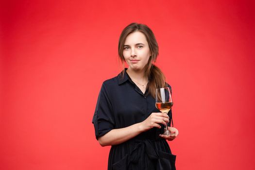 Young pretty lady in casual clothes holding delicious drink in hands. Brunette happy girl enjoying her sweet beverage and smiling. Beautiful woman in black dress standing with glass of expensive wine.