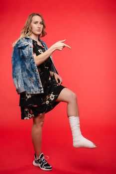 Side view of girl standing on red isolated background with broken leg. Young womab in dress and denim jacken pointing with finger at fracture. Concept of cast and gypsum.