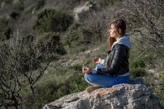 Woman tourist enjoying the sunset over the sea mountain landscape. Sits outdoors on a rock above the sea. She is wearing jeans, a blue hoodie and a black leather jacket. Healthy lifestyle, harmony and meditation.
