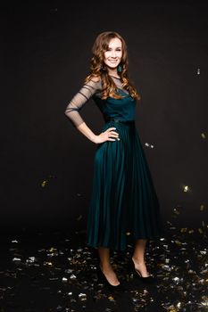 Happy young girl looking surprisingly while golden confetti falling. Beautiful woman in emerald elegant long dress smiling and dancing among decorations. Gorgeous lady celebrating birthday on party.