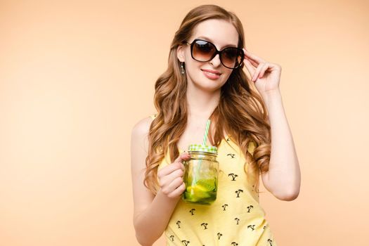 Young happy lady standing on one leg and holding other one up. Brunette girl in casual clothes enjoying tasty fruit cocktail. Beautiful woman in sunglasses holding jar with citrus lemonade in hands.