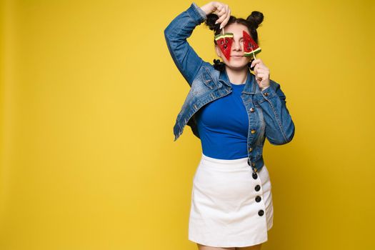 Full length studio portrait of laughing model in white top and green skirt and heels holding two sweet candies on her eyes like sunglasses with her mouth open. Isolate on yellow.