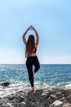 Young woman in black leggings and black top with long flowing hair practicing stretching outdoors by the sea on a sunny day. Women's yoga, fitness, pilates. The concept of a healthy lifestyle, harmony