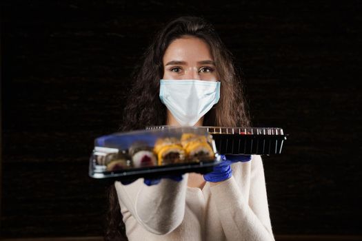 Girl sushi courier in medical mask with 2 boxes. Sushi set in box healthy food delivery service by car. Japanese cuisine: rolls, soy sauce, wasabi.