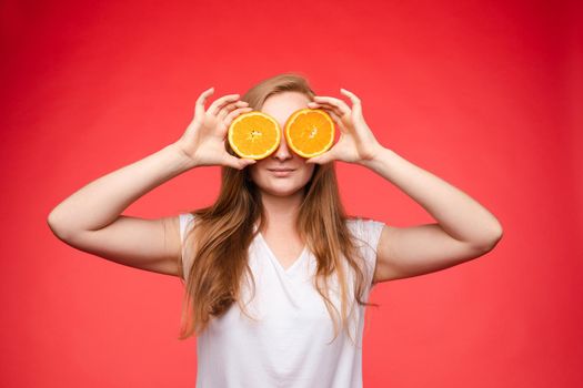 Studio headshot of young funny brunette with hairstyle and red lips holding halved oranges on eyes against bright yellow background.