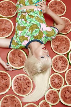 View from above fashionable blonde with makeup lying near fresh tasty watermelon. Attractive female model looking at camera and posing with sweet fruits. Concept of summer and brightness.