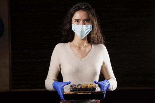 Girl with sushi. Food courier in medical mask. Delivery at quarantine coronavirus covid-19. Young woman holding 2 sushi boxes in hands using gloves