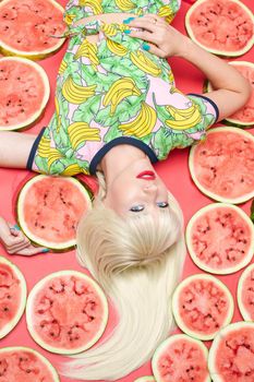 View from above fashionable blonde with makeup lying near fresh tasty watermelon. Attractive female model looking at camera and posing with sweet fruits. Concept of summer and brightness.