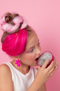 Close-up portrait of pretty girl with pink hairstyle with dragon fruit on pink background. Studio shot of charming tween girl with pink make up enjoying juicy red pitaya. exotic Pitahaya fruit.