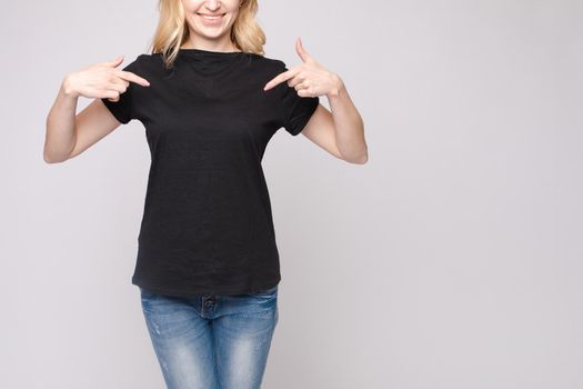 Cropped view from front of happy blond standing in black shirt and jeans and pointing at outfit with fingers. Cheerful girl laughing and posing on grey isolated backround. Concept of casual style.