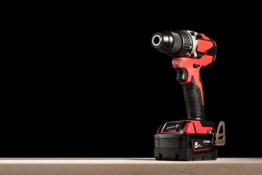 a cordless screwdriver stands on a wooden table on a black background. Cordless drill with lithium-ion battery in red. Professional tool for drilling holes and driving screws