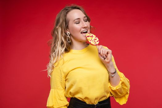 Studio portrait of attractive young woman with long wave hair enjoying tasty sweet colorful lollipop with her eyes closed. She is wearing bright yellow blouse and fashionable earrings. Isolate on red.