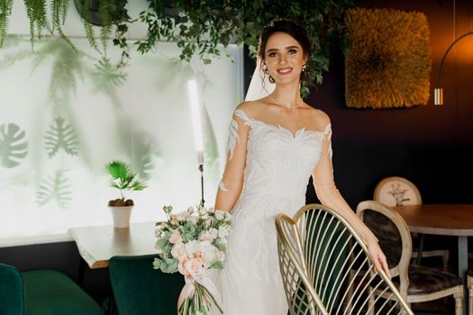Bride with wedding bouquet smiles, looks in camera and touches her face. Attractive girl portrait for social networks. Girl in wedding dress in luxury restaurant