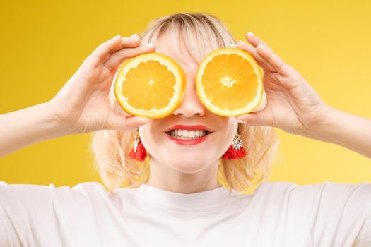 Front view of cheerful young blonde posing with fresh oranges on isolated background. Funny girl keeping fruit and closing eyes in studio. Concept of happiness and health.