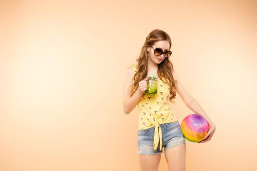 Side view of sporty blonde drinking fresh juice, doing sport and posing in studio. Young model keeping ball and racket and smiling on isolated background. Concept of energy and power.