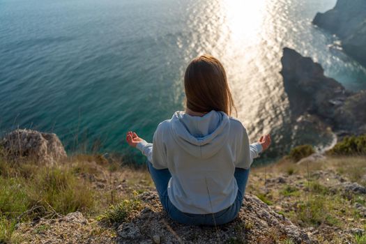 Woman tourist enjoying the sunset over the sea mountain landscape. Sits outdoors on a rock above the sea. She is wearing jeans and a blue hoodie. Healthy lifestyle, harmony and meditation.