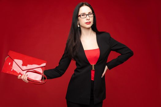 Studio portrait of stylish elegant business woman in glasses and black suit holding a trendy glossy handbag. She is isolated on bright red background.