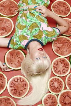 View from above fashionable blonde with makeup lying near fresh tasty watermelon. Attractive female model looking at camera and posing with sweet fruits. Concept of summer and brightness.