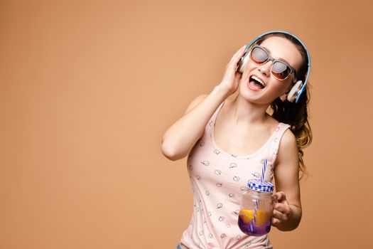 Attractive young girl in big sunglasses singing favourite songs during free time. Beautiful woman in blue headphones listening music and smiling. Brunette lady relaxing and drinking tasty lemonade.