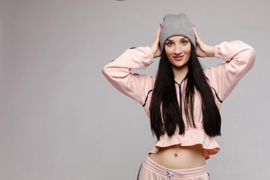 Beautiful girl with big blue eyes and opened belly posing at camera. Brunette happy girl with long hair in pink costume smiling. Stylish model in sportswear and grey hat holding head by both hands.