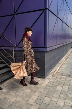 A happy shopaholic girl keeps her bags near the shopping center. A woman near the store is happy with her purchases, holding bags. Dressed in a leopard print dress. Consumer concept