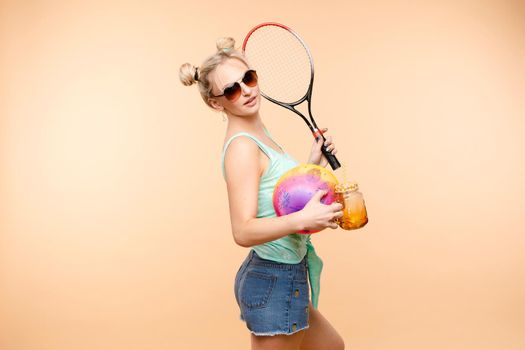 Side view of sporty blonde drinking fresh juice, doing sport and posing in studio. Young model keeping ball and racket and smiling on isolated background. Concept of energy and power.