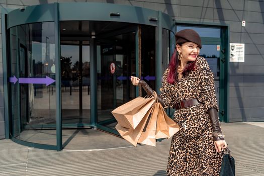 A happy shopaholic girl keeps her bags near the shopping center. A woman near the store is happy with her purchases, holding bags. Dressed in a leopard print dress. Consumer concept