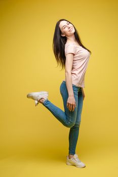 Full length isolate portrait of positive brunette young girl in jeans and t-shirt and sneakers jumping over yellow background. She is smiling at camera.