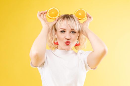 Beautiful model holding an orange. Smiles, makes sponge. Positive photo. Bright makeup. Isolated on yellow background.