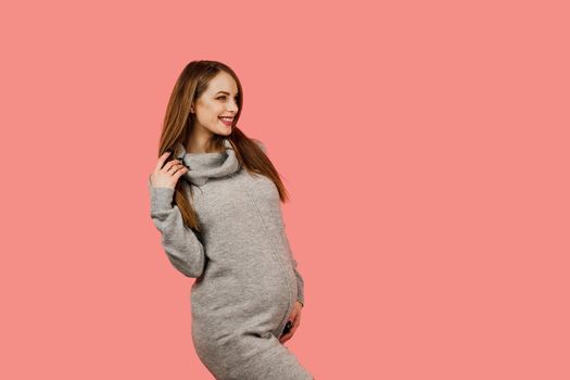 Pregnant woman. Expectation of the child. Maternity leave. Pregnancy of a happy beautiful girl in a gray sweater on a pink background.