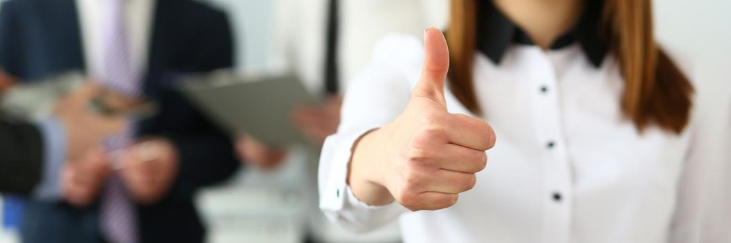 Focus on woman hand showing thumb at camera. Smiling female standing near witty colleagues discussing questionable subject of business in modern building. Company meeting concept. Blurred background