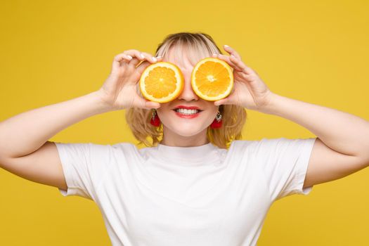 Beautiful model holding an orange. Smiles, makes sponge. Positive photo. Bright makeup. Isolated on yellow background.