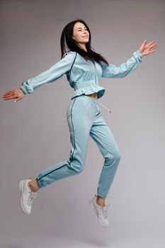 Happy brunette wearing blue tracksuit jumping and laughing on grey isolated background. Young cheerful woman with long hair having fun and posing in studio. Concept of happiness and motion.