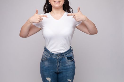 Cropped view from front of happy brunette standing in white shirt and jeans and pointing at outfit with fingers. Cheerful girl laughing and posing on grey isolated backround. Concept of casual style.