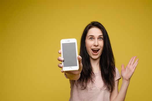 Studio portrait of amazed young caucasian woman with wavy hair wearing white top and green bottom holding mobile phone screen to camera. She is shocked or surprised with the news or info on the screen. Copyspace. Isolate on yellow.