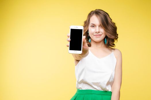 Studio portrait of amazed young caucasian woman with wavy hair wearing white top and green bottom holding mobile phone screen to camera. She is shocked or surprised with the news or info on the screen. Copyspace. Isolate on yellow.