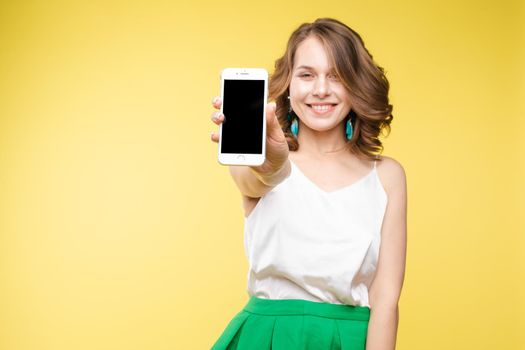 Studio portrait of amazed young caucasian woman with wavy hair wearing white top and green bottom holding mobile phone screen to camera. She is shocked or surprised with the news or info on the screen. Copyspace. Isolate on yellow.