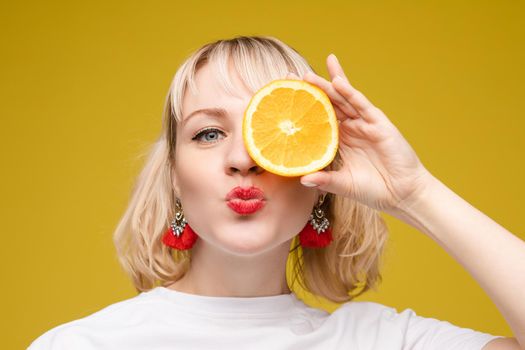 Studio headshot of young funny brunette with hairstyle and red lips holding halved oranges on eyes against bright yellow background.