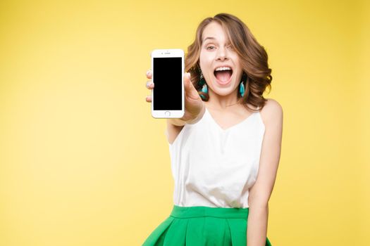 Studio portrait of amazed young caucasian woman with wavy hair wearing white top and green bottom holding mobile phone screen to camera. She is shocked or surprised with the news or info on the screen. Copyspace. Isolate on yellow.