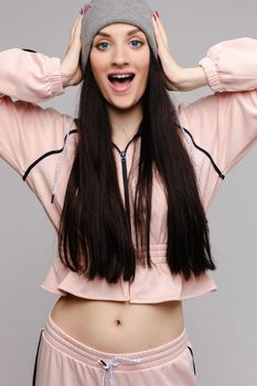 Beautiful girl with big blue eyes and opened belly posing at camera. Brunette happy girl with long hair in pink costume smiling. Stylish model in sportswear and grey hat holding head by both hands.