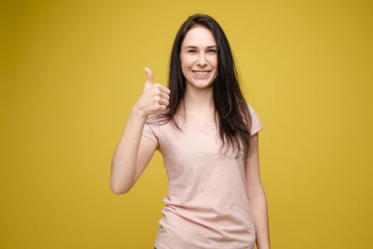 Front view of positive blonde wearing white shirt and jeans looking at camera and showing thumbs up. Young woman posing and laughing on grey isolated background. Concept of happiness.