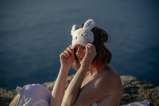 Young woman in sleep mask posing on the seashore. She is wrapped in a white blanket. The coast of the sea at a height, around the sea and rocks, dawn