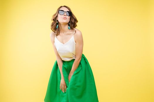 Summer outfit on orange background. Stylish and trendy beautiful young girl with wavy middle-length hair in white top and green bottom wearing trendy earrings and black sunglasses. She is smiling over orange background. Copyspace. Isolate.