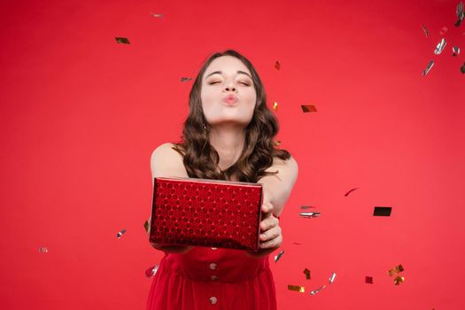 Front view of pretty brunette wearing red dress looking at camera while sparkles falling around in studio. Beautiful woman with hairstyle posing on isolated background. Concept of holiday.